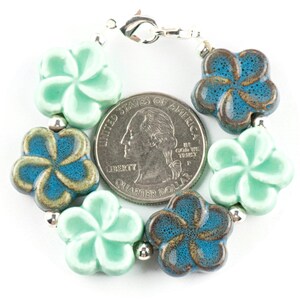May include: A bracelet made of ceramic flower beads in shades of teal and green. The beads are in the shape of a plumeria flower. The bracelet has a silver clasp.