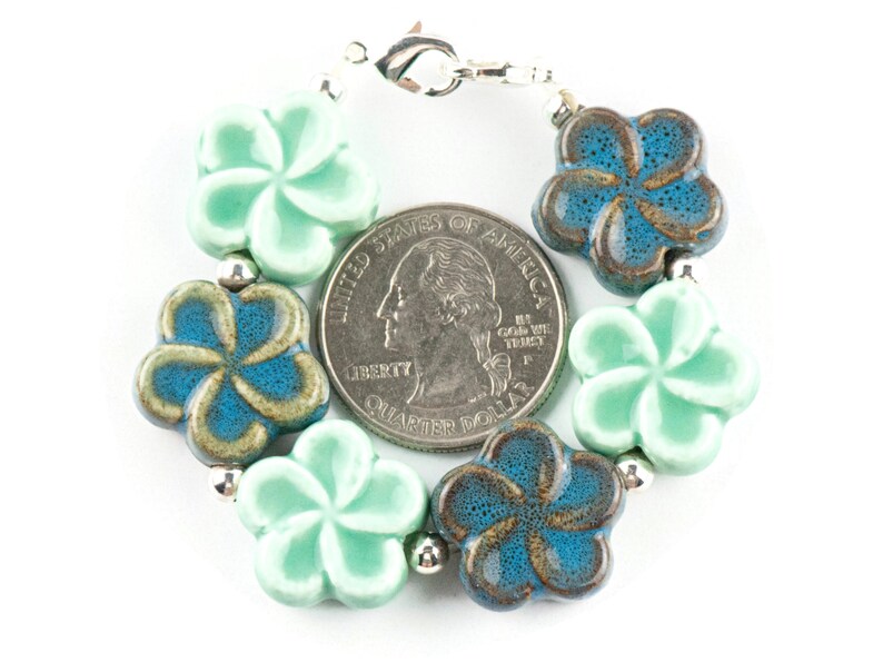 May include: A bracelet made of ceramic flower beads in shades of teal and green. The beads are in the shape of a plumeria flower. The bracelet has a silver clasp.