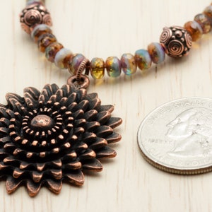 Puede incluir: Un collar de cobre con un gran colgante de girasol de cobre. El collar est&aacute; hecho con cuentas de vidrio iridiscente y alambre de cobre. El colgante es aproximadamente del tama&ntilde;o de una moneda de 25 centavos.