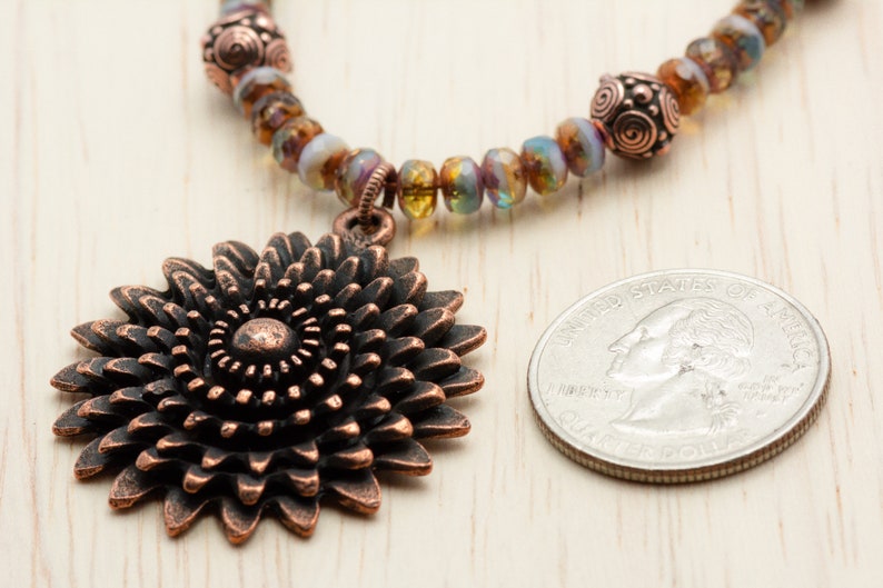 Puede incluir: Un collar de cobre con un gran colgante de girasol de cobre. El collar est&aacute; hecho con cuentas de vidrio iridiscente y alambre de cobre. El colgante es aproximadamente del tama&ntilde;o de una moneda de 25 centavos.