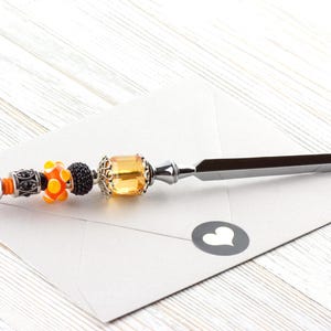 May include: A decorative letter opener with a silver blade and a beaded handle. The handle features orange, black, and yellow beads, and a silver end. The letter opener rests on a white envelope with a heart-shaped sticker.