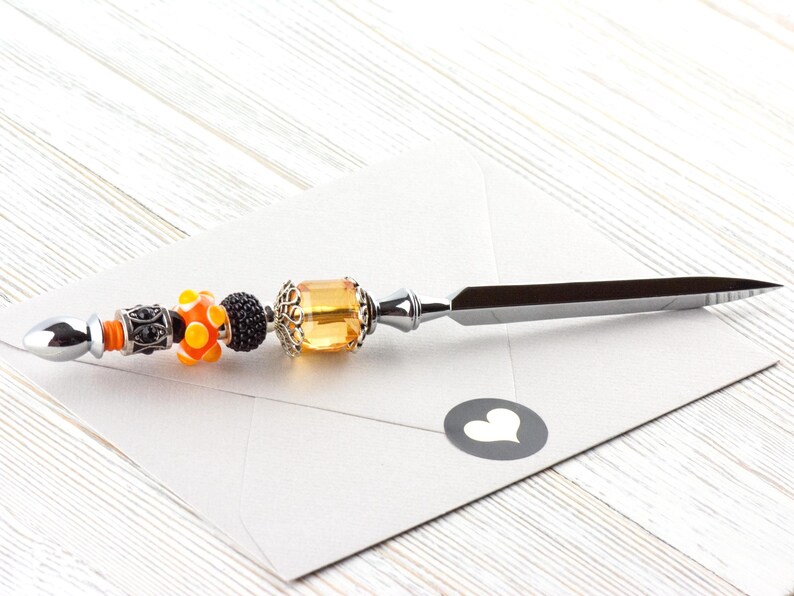 May include: A decorative letter opener with a silver blade and a beaded handle. The handle features orange, black, and yellow beads, and a silver end. The letter opener rests on a white envelope with a heart-shaped sticker.