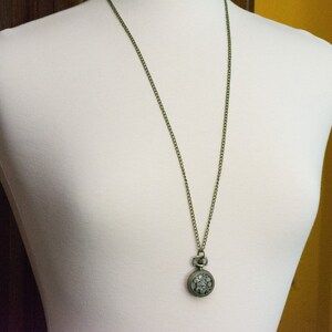 May include: A silver pocket watch with a detailed design hangs from a bronze chain on a white mannequin.
