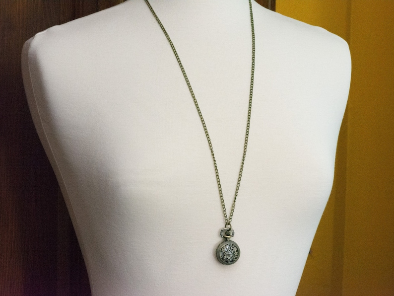 May include: A silver pocket watch with a detailed design hangs from a bronze chain on a white mannequin.