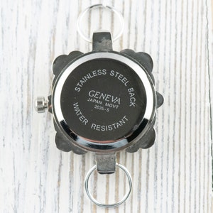 May include: The back of a silver watch with a square face. The watch has a silver band and a silver clasp. The watch face has the words "Stainless Steel Back" and "Geneva Japan Movt 2035-S Water Resistant" printed on it.