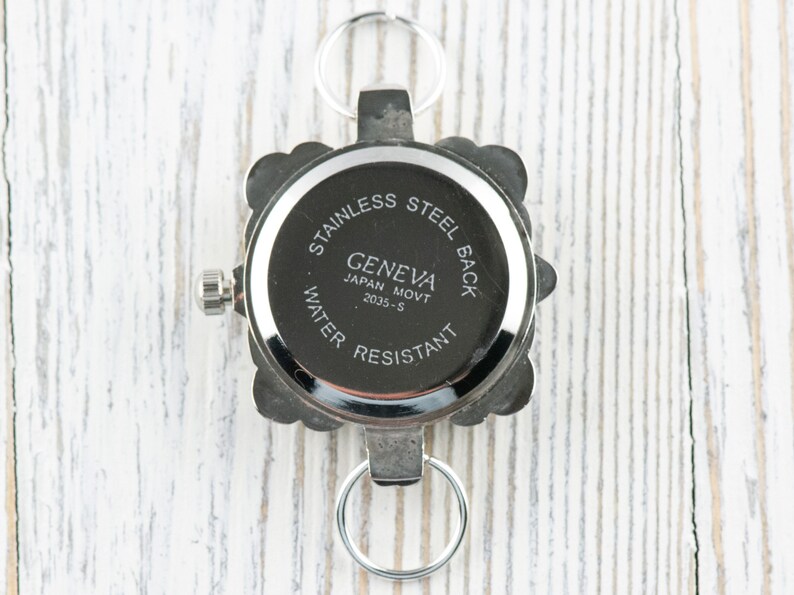 May include: The back of a silver watch with a square face. The watch has a silver band and a silver clasp. The watch face has the words "Stainless Steel Back" and "Geneva Japan Movt 2035-S Water Resistant" printed on it.
