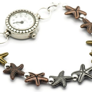 May include: A silver watch with a white face and black hands is attached to a bracelet with silver and bronze starfish charms.