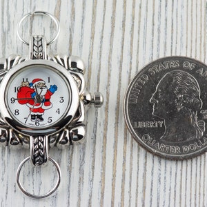 May include: A silver-toned pocket watch with a white face featuring a cartoon Santa Claus holding a bag of gifts. The watch is about the size of a US quarter.