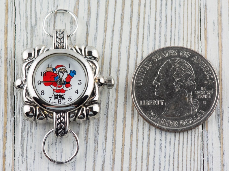 May include: A silver-toned pocket watch with a white face featuring a cartoon Santa Claus holding a bag of gifts. The watch is about the size of a US quarter.