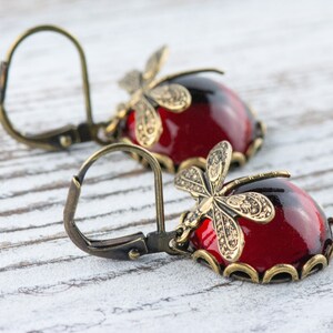 Pode incluir: Um par de brincos vermelhos e dourados com detalhes de lib&eacute;lula. Os brincos t&ecirc;m um caboch&atilde;o de vidro vermelho em forma de l&aacute;grima com um design de lib&eacute;lula dourada na parte superior. Os brincos s&atilde;o presos a um gancho dourado.