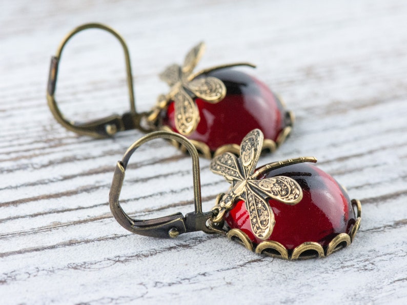 Pode incluir: Um par de brincos vermelhos e dourados com detalhes de lib&eacute;lula. Os brincos t&ecirc;m um caboch&atilde;o de vidro vermelho em forma de l&aacute;grima com um design de lib&eacute;lula dourada na parte superior. Os brincos s&atilde;o presos a um gancho dourado.