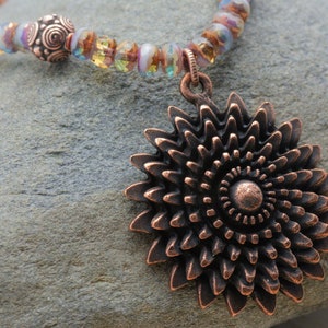 Puede incluir: Un collar de cobre con un gran colgante en forma de flor intrincado. El collar est&aacute; hecho con cuentas de vidrio iridiscente y cuentas de cobre.