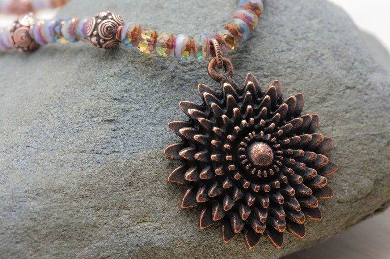 Puede incluir: Un collar de cobre con un gran colgante en forma de flor intrincado. El collar est&aacute; hecho con cuentas de vidrio iridiscente y cuentas de cobre.