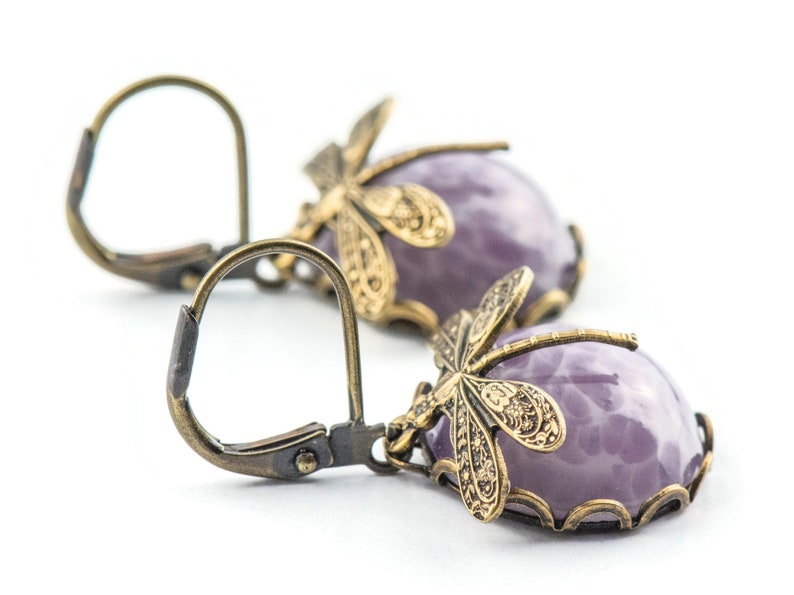 Pode incluir: Um par de brincos pendentes com uma pedra roxa e um design de lib&eacute;lula. Os brincos s&atilde;o feitos de lat&atilde;o e t&ecirc;m um fecho de gancho.