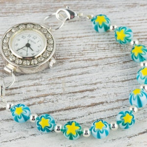 Peut inclure: Une montre argent&eacute;e avec un cadran blanc et une lunette perl&eacute;e est attach&eacute;e &agrave; un bracelet compos&eacute; de perles argent&eacute;es et de perles de verre bleues et blanches avec des motifs d'&eacute;toiles jaunes.