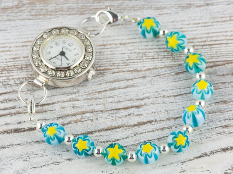Peut inclure: Une montre argent&eacute;e avec un cadran blanc et une lunette perl&eacute;e est attach&eacute;e &agrave; un bracelet compos&eacute; de perles argent&eacute;es et de perles de verre bleues et blanches avec des motifs d'&eacute;toiles jaunes.