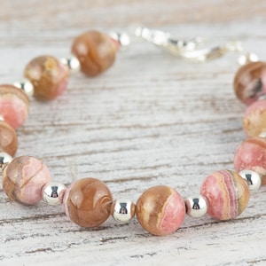 May include: A pink and brown gemstone bracelet with silver beads. The bracelet is made with round, polished stones and has a silver clasp.