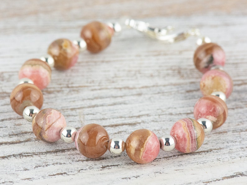 May include: A pink and brown gemstone bracelet with silver beads. The bracelet is made with round, polished stones and has a silver clasp.