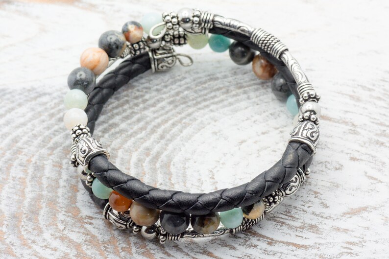 May include: A black leather bracelet with silver beads and charms. The bracelet features a variety of stones, including green, brown, and white.