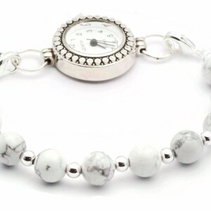 May include: A silver watch bracelet with a white watch face and a beaded band. The band features white, marbled beads interspersed with small silver beads. The watch face is round with black numerals and hands.