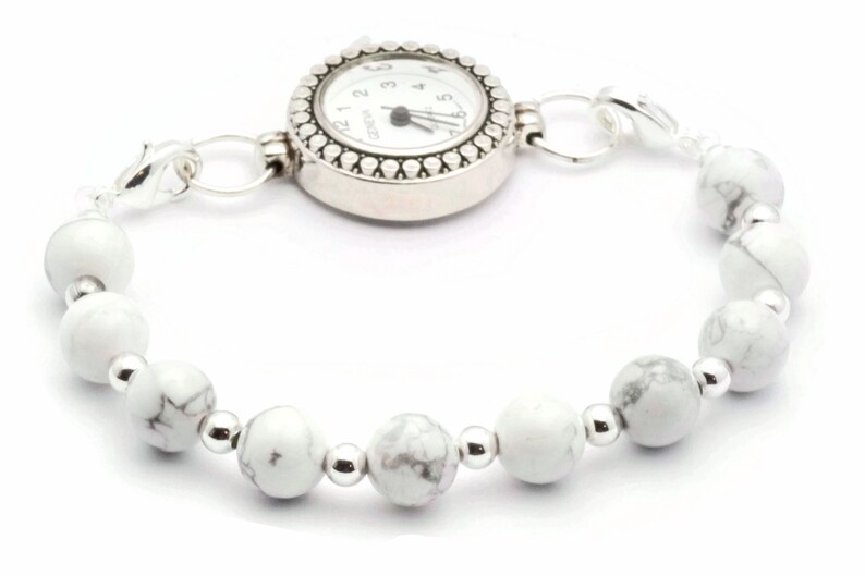 May include: A silver watch bracelet with a white watch face and a beaded band. The band features white, marbled beads interspersed with small silver beads. The watch face is round with black numerals and hands.