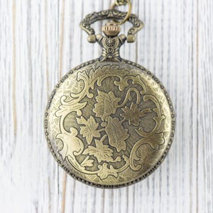 May include: A gold-toned pocket watch with an ornate floral design on the front. The watch has a chain attached to the top.