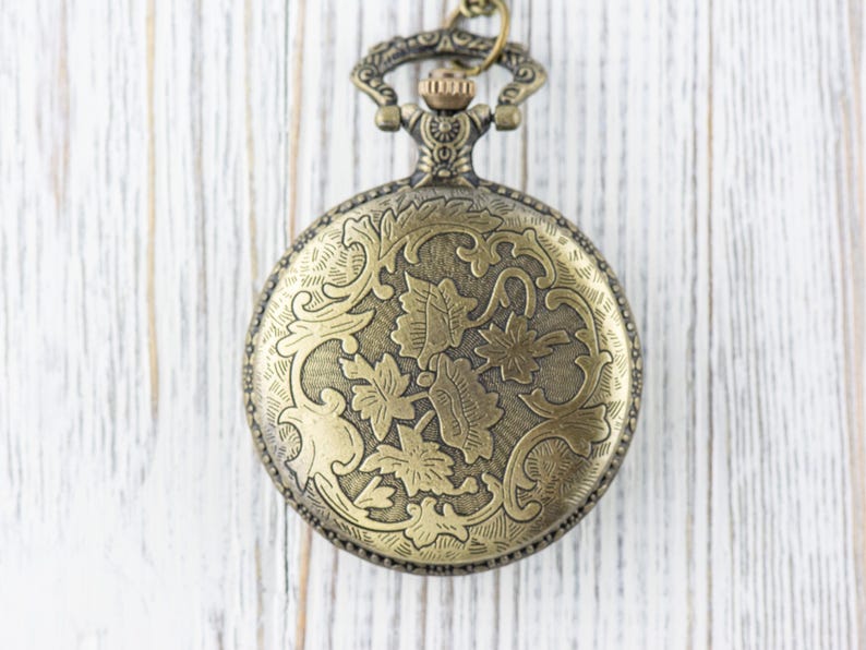 May include: A gold-toned pocket watch with an ornate floral design on the front. The watch has a chain attached to the top.