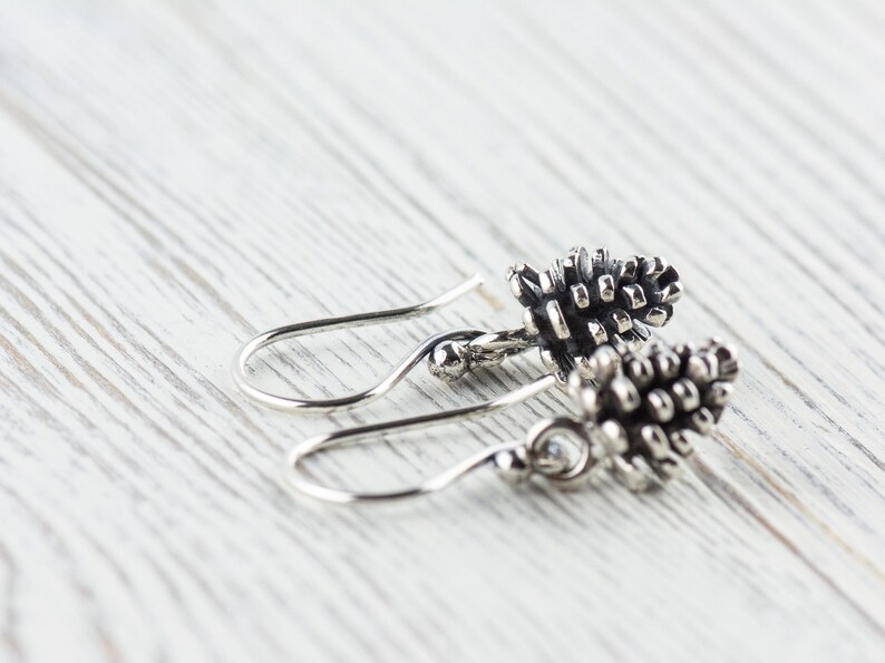 May include: A pair of silver earrings with a pine cone design. The earrings are dangle style with a small silver bead at the top of the hook.