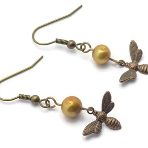 May include: A pair of gold-toned earrings featuring a small bee charm and a golden pearl bead on each earring.