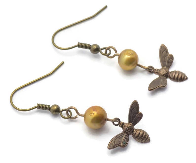 May include: A pair of gold-toned earrings featuring a small bee charm and a golden pearl bead on each earring.