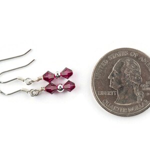 Puede incluir: Un par de pendientes de plata con dos cuentas de cristal facetado de color rojo oscuro en cada pendiente. Los pendientes se muestran junto a una moneda de veinticinco centavos de d&oacute;lar estadounidense para comparar el tama&ntilde;o.