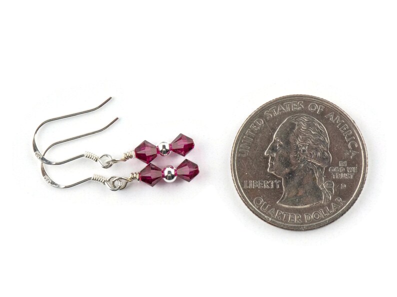 Puede incluir: Un par de pendientes de plata con dos cuentas de cristal facetado de color rojo oscuro en cada pendiente. Los pendientes se muestran junto a una moneda de veinticinco centavos de d&oacute;lar estadounidense para comparar el tama&ntilde;o.