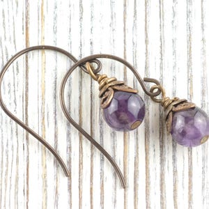 K&ouml;nnte beinhalten: Handgefertigte Ohrringe mit tropfenf&ouml;rmigen Amethyst-Edelsteinen. Die violetten Steine h&auml;ngen an bronzefarbenen Ohrhaken. Die Ohrringe haben einen Vintage- und Boh&egrave;me-Stil, ideal f&uuml;r den Alltag oder besondere Anl&auml;sse.