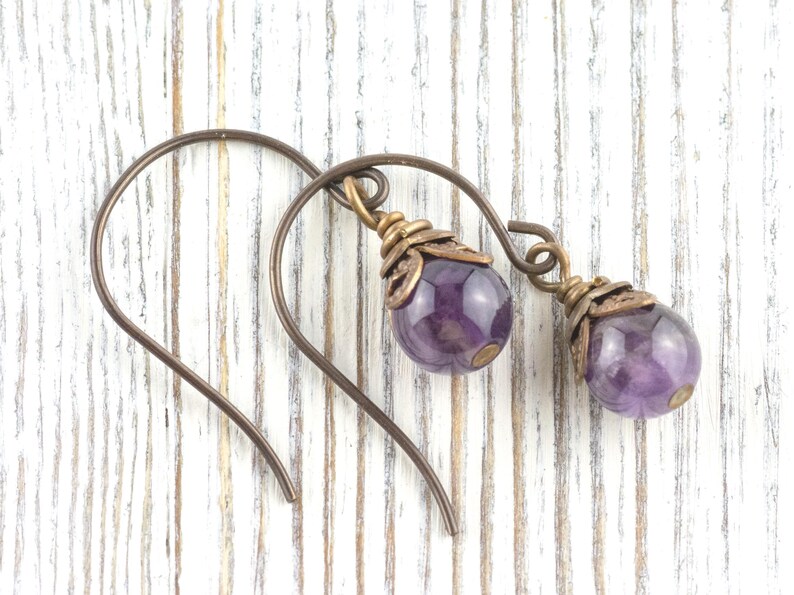 K&ouml;nnte beinhalten: Handgefertigte Ohrringe mit tropfenf&ouml;rmigen Amethyst-Edelsteinen. Die violetten Steine h&auml;ngen an bronzefarbenen Ohrhaken. Die Ohrringe haben einen Vintage- und Boh&egrave;me-Stil, ideal f&uuml;r den Alltag oder besondere Anl&auml;sse.