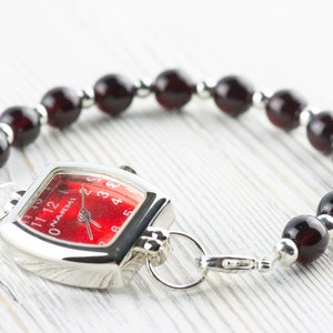May include: A silver watch with a red face and black Roman numeral hour markers. The watch is attached to a bracelet made of dark red beads and silver spacers.