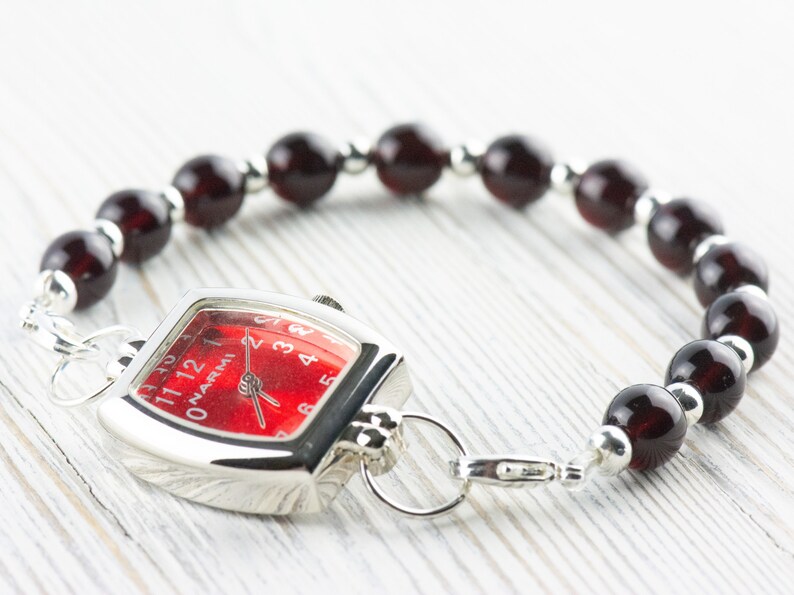 May include: A silver watch with a red face and black Roman numeral hour markers. The watch is attached to a bracelet made of dark red beads and silver spacers.