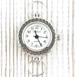May include: Silver Geneva quartz watch with a white face and black hands. The watch has a circular design with a decorative bezel and two attached rings. The watch face displays the time with black numerals and the word "GENEVA".