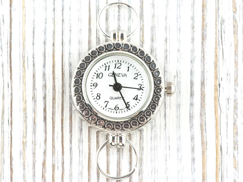 May include: Silver Geneva quartz watch with a white face and black hands. The watch has a circular design with a decorative bezel and two attached rings. The watch face displays the time with black numerals and the word "GENEVA".