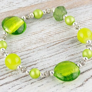 Puede incluir: Un brazalete de cuentas de vidrio verde con detalles plateados. El brazalete presenta una variedad de cuentas de vidrio verde, incluyendo formas redondas, ovaladas e irregulares.