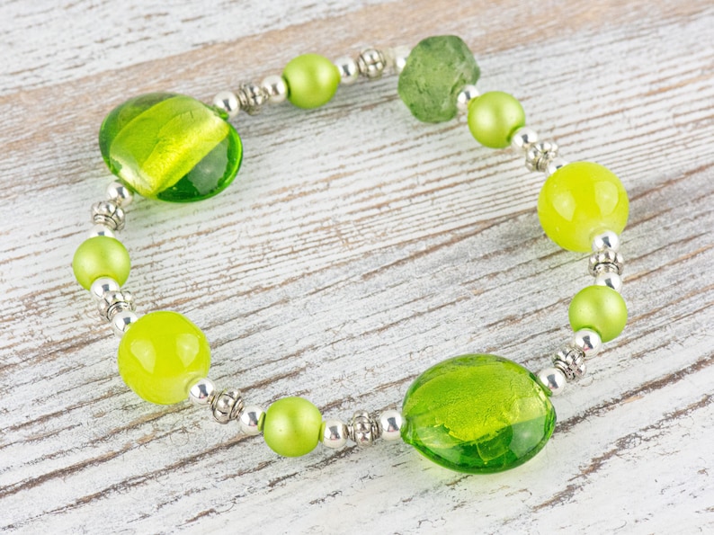 Puede incluir: Un brazalete de cuentas de vidrio verde con detalles plateados. El brazalete presenta una variedad de cuentas de vidrio verde, incluyendo formas redondas, ovaladas e irregulares.