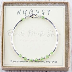 Op de afbeelding: Een zilveren armband met groene peridot kristallen en het woord "AUGUST" op een witte kaart in een bruine doos.