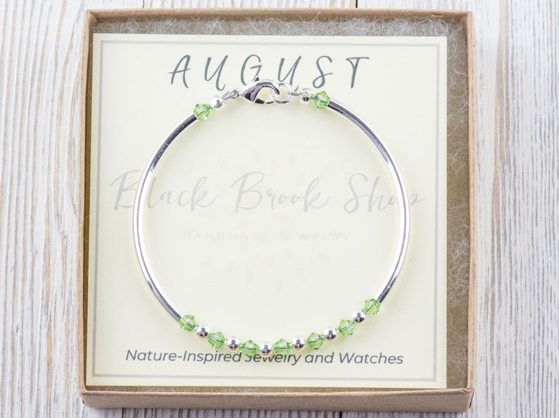 Op de afbeelding: Een zilveren armband met groene peridot kristallen en het woord "AUGUST" op een witte kaart in een bruine doos.