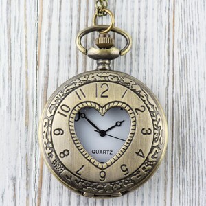 Pode incluir: Um rel&oacute;gio de bolso dourado com uma janela em forma de cora&ccedil;&atilde;o que revela um mostrador branco com ponteiros pretos e a palavra "QUARTZ". O rel&oacute;gio tem numerais romanos e um design intrincado ao redor da borda.
