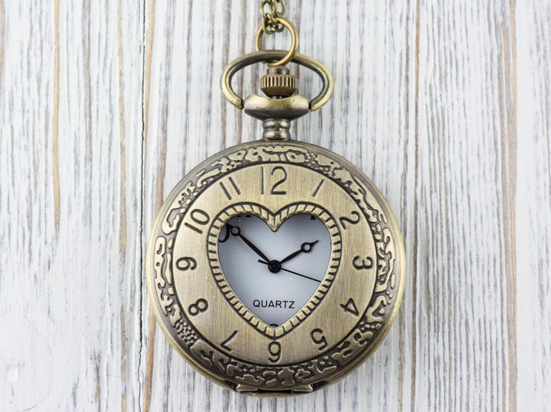 Pode incluir: Um rel&oacute;gio de bolso dourado com uma janela em forma de cora&ccedil;&atilde;o que revela um mostrador branco com ponteiros pretos e a palavra "QUARTZ". O rel&oacute;gio tem numerais romanos e um design intrincado ao redor da borda.