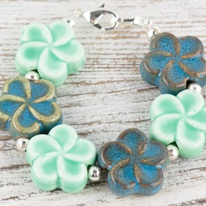 May include: A bracelet made of ceramic flower-shaped beads in shades of blue and green. The bracelet is strung on a silver chain.