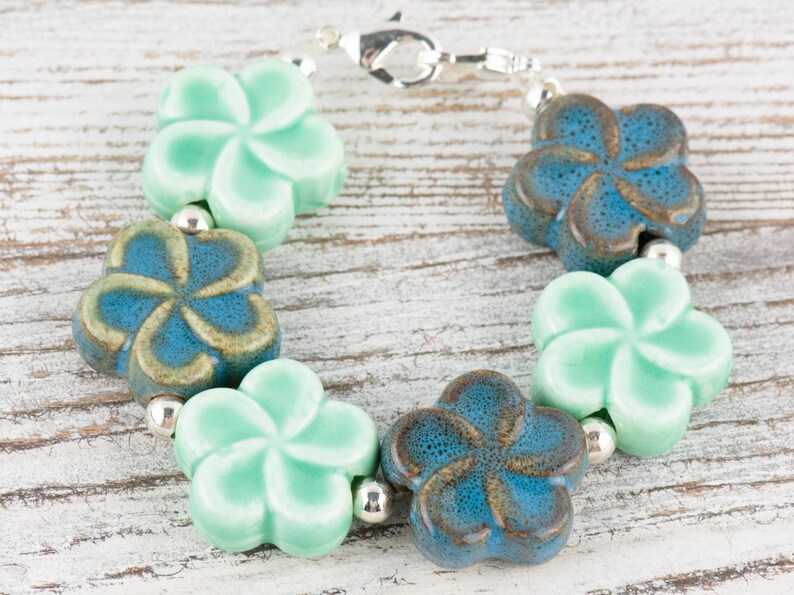 May include: A bracelet made of ceramic flower-shaped beads in shades of blue and green. The bracelet is strung on a silver chain.