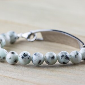 K&ouml;nnte beinhalten: Ein silbernes Armband mit einem Design aus wei&szlig;en und schwarzen, gefleckten Steinperlen. Das Armband hat ein silbernes Schild mit einem leeren Platz zum Gravieren.