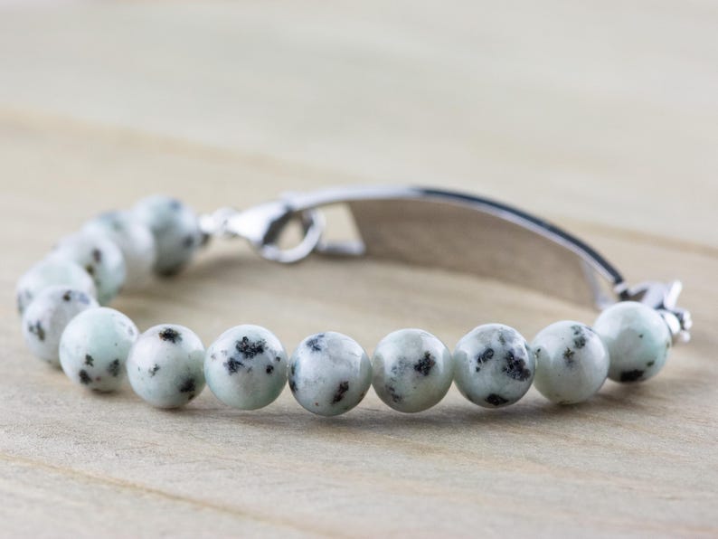 K&ouml;nnte beinhalten: Ein silbernes Armband mit einem Design aus wei&szlig;en und schwarzen, gefleckten Steinperlen. Das Armband hat ein silbernes Schild mit einem leeren Platz zum Gravieren.
