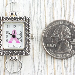 May include: Silver-toned rectangular watch with a pink ribbon design on the face, next to a U.S. quarter. The watch face displays the time and the word "GENEVA". The watch has a decorative frame and two ring attachments.