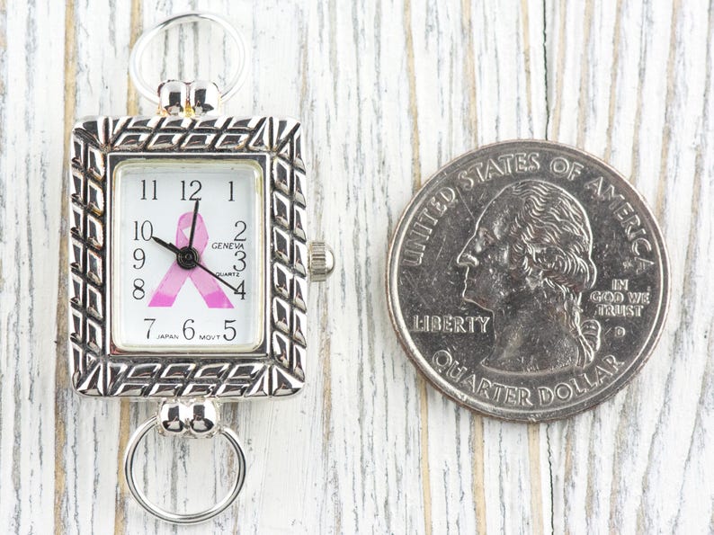 May include: Silver-toned rectangular watch with a pink ribbon design on the face, next to a U.S. quarter. The watch face displays the time and the word "GENEVA". The watch has a decorative frame and two ring attachments.